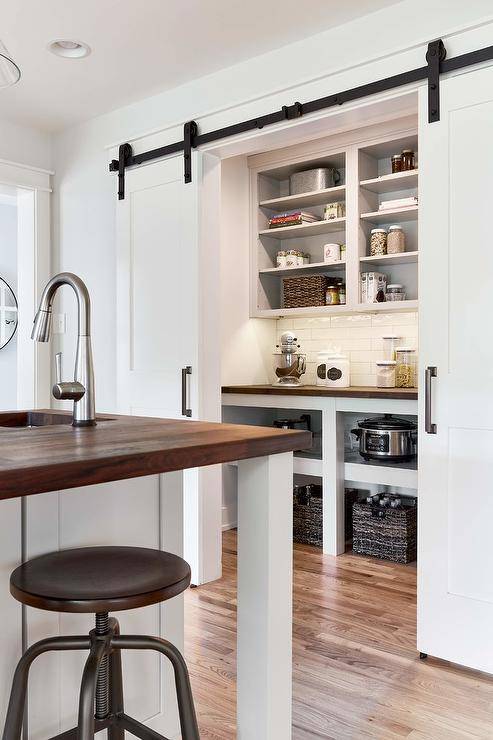 Dual white doors on oil rubbed bronze rails open to a small walk in pantry boasting white shelves positioned above a wood countertop mounted over shelves and against white staggered wall tiles.