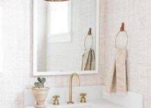 A tan washstand fitted with a gold gooseneck faucet is positioned benaeth a white framed vanity mirror hung from a wall clad in tan textured walllpaper beneath a Cerro Wall Sconce.