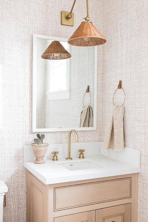 A tan washstand fitted with a gold gooseneck faucet is positioned benaeth a white framed vanity mirror hung from a wall clad in tan textured walllpaper beneath a Cerro Wall Sconce.