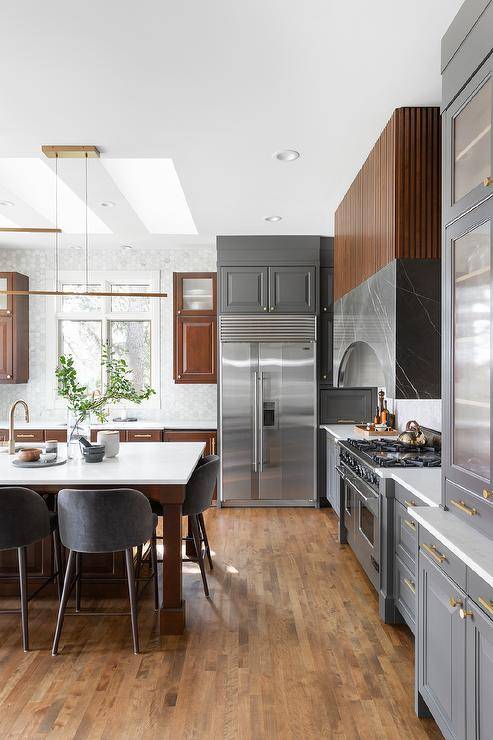Kitchen features a brown reeded and gray marble hood over a cooktop and gray velvet dining chairs at a brown wooden center island with sink that boasts a brass gooseneck faucet illuminated by staggered linear island lights and gray and brown two tone kitchen cabinetry.