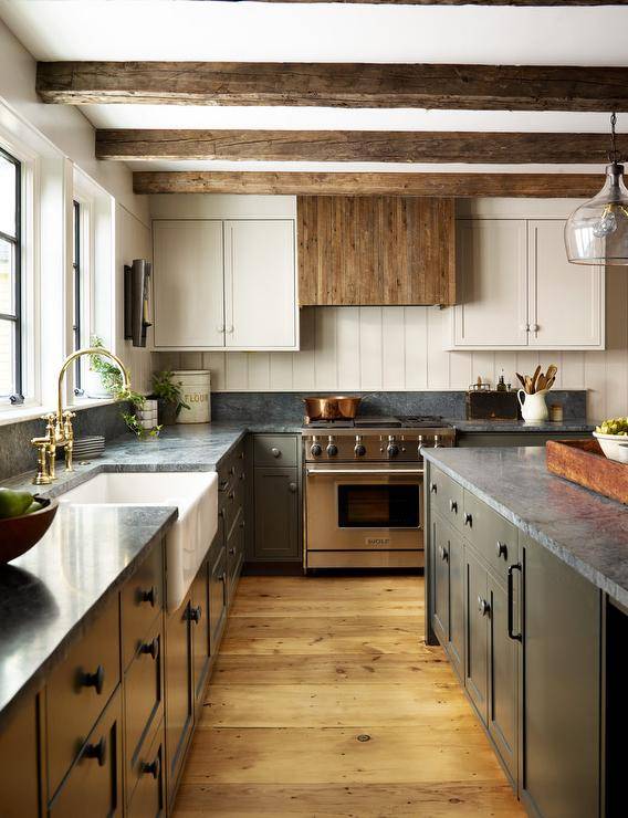 Kitchen features a reclaimed wood hood over a Wolf range with shiplap trim, two tone kitchen cabinets topped with gray marble and an apron sink with brass gooseneck faucet under wooden ceiling beams.