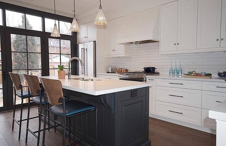 Two-tone vintage island stools at a black center island under antiqued mercury glass lights pendants. White quartz countertops balance the black island while the perimiter space is equipped with horizontal white tiles, a white vent hood, and chrome hardware.