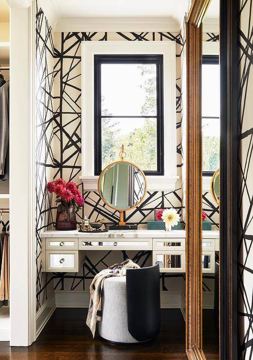 Makeup vanity nook with Kelly Wearstler Channels wallpaper features a gray fabric and black leather vanity stools at a mirrored makeup vanity under a window.