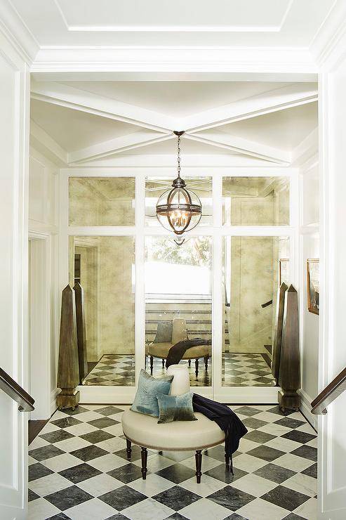 Stunning black and white foyer is illuminated by a Restoration hardware Victorian Hotel Pendant hung from a white ceiling accented with an x-trim over a round ivory leather ottoman placed on white and black harlequin floor tiles in front of a mirrored paneled wall.