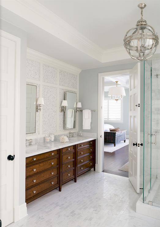 Chic master bathroom features a Restoration Hardware Victorian Hotel Pendant illuminating a seamless glass shower clad in marble. A linear marble tiled floor leads to a stained double washstand topped with white marble fitted with his and hers sinks placed under polished nickel pivot mirrors lining a mosaic marble tiled wall.