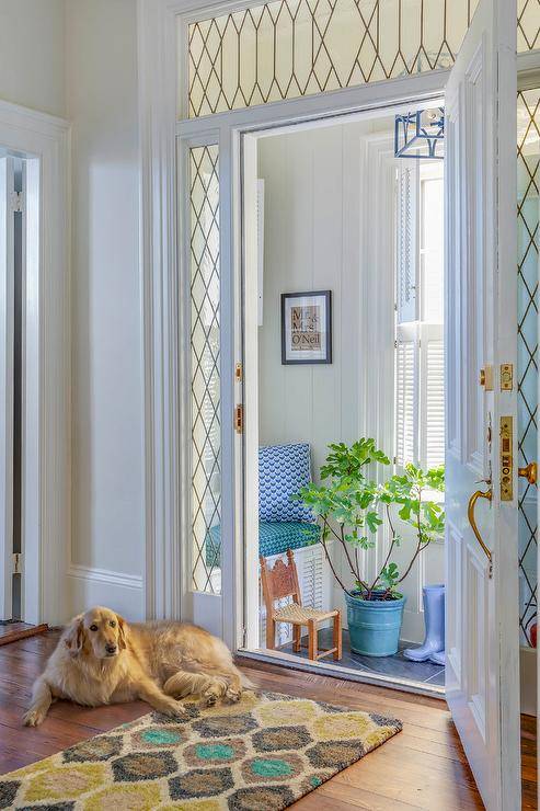 Leaded sidelights and a leaded glass transom window frame a white door open to a long and narrow vestibule boasting a white built-in bench topped with a turquoise blue cushion accented with a white and blue pillow placed against a shiplap wall.