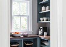 A brass semi-flush mount lights a small pantry fitted with gray blue open cabinets topped with a butcher block countertop fixed beneath a window and gray-blue shelves. Walls are lined with gray-blue crown moldings.