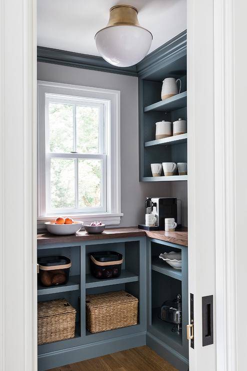 A brass semi-flush mount lights a small walk in pantry fitted with gray blue open cabinets topped with a butcher block countertop fixed beneath a window and gray-blue shelves. Walls are lined with gray-blue crown moldings.