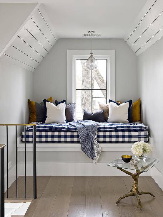 Attic window seat nook boasts a blue buffalo check cushion with various accent pillows and a gold faux bios accent table in a cottage home.