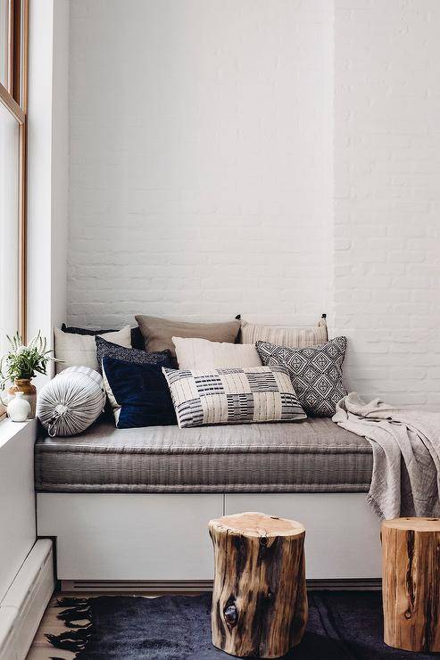 Window seat bench with gray cushions is next to tree trunk accent tables on a black fringe rug.