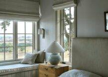 Bedroom features a built in paneled window seat and a glossy white lamp that illuminates a vintage oak nightstand with an oatmeal linen headboard under an ivory plank vaulted ceiling.