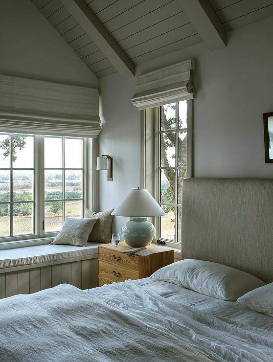 Bedroom features a built in paneled window seat and a glossy white lamp that illuminates a vintage oak nightstand with an oatmeal linen headboard under an ivory plank vaulted ceiling.