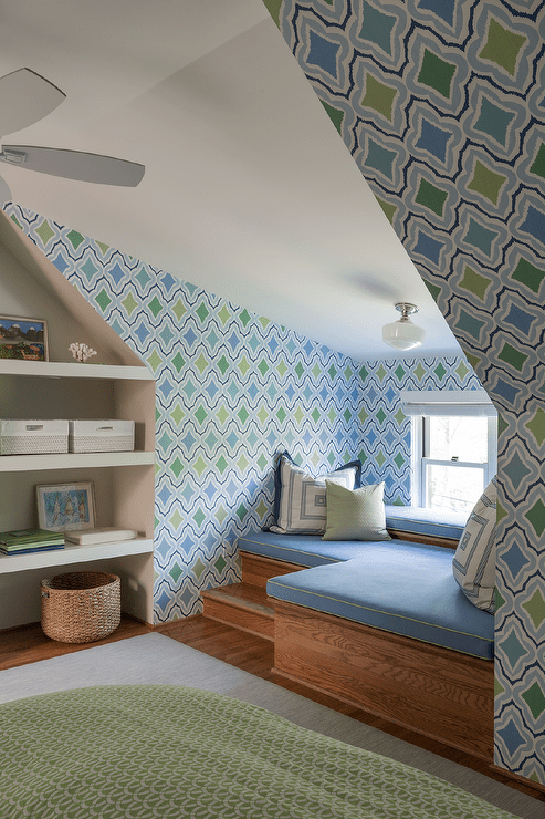 L shaped built in window seat with green and blue geometric wallpaper features blue cushions next to a built in bookshelf with green rug.