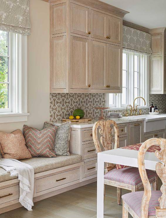 Welcoming kitchen features a brushed oak built-in window seat donning drawers with brass pulls and topped with a tan seat cushion complemented with an assortment of pillows.