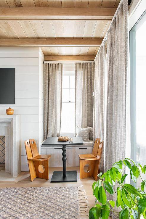 Gray curtains hang over a gray built-in window seat topped with gray cushions and pillow and fitted with storage drawers. Brown wood chairs flank a black table placed in front of the window seat and beneath a tan wood plank ceiling.