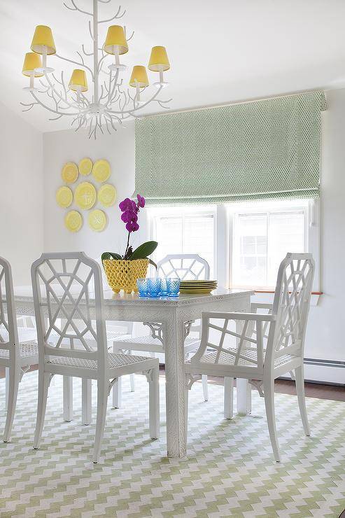 White trellis chairs and a white faux bois dining table in a dining room completed with a green trellis rug, a white faux bois chandelier and yellow plate wall decor. A green roman shade complements the dining room accents while contrasting the white walls and furnishings.