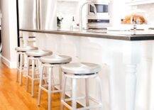 Crate & Barrel Spin Swivel Backless Counter Stools are accessible and chic. Black and white kitchen island features turned legs at the ends with corbels adding a luxurious touch. Black quartz countertops contrast the all white kitchen decor while complementing steel appliances and silver accents.