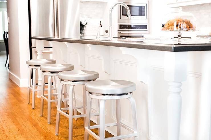 Crate & Barrel Spin Swivel Backless Counter Stools are accessible and chic. Black and white kitchen island features turned legs at the ends with corbels adding a luxurious touch. Black quartz countertops contrast the all white kitchen decor while complementing steel appliances and silver accents.