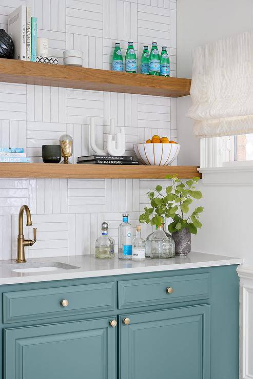 Blue kitchenette cabinetry boasts an antique brass gooseneck faucet fixed beneath stacked floating shelves mounted to white basketweave wall tiles.