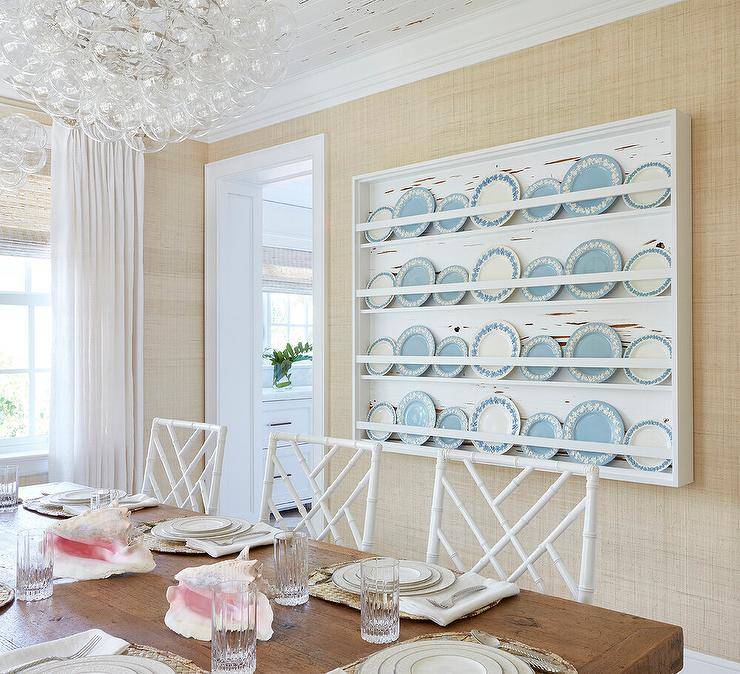 White bamboo lattice dining chairs sit at a reclaimed wood dining table lit by a glass bubbles chandelier and in front of a white plate display rack fixed to a wall covered in beige wallpaper.