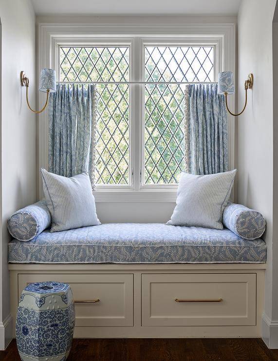 Blue café curtains cover leaded windows positioned over a built-in window seat fitted with drawers donning brass pulls and a white and blue cushion complemented with white and blue lumbar pillows and blue striped pillows. The window seat is lit by facing Reed Single Sconces.