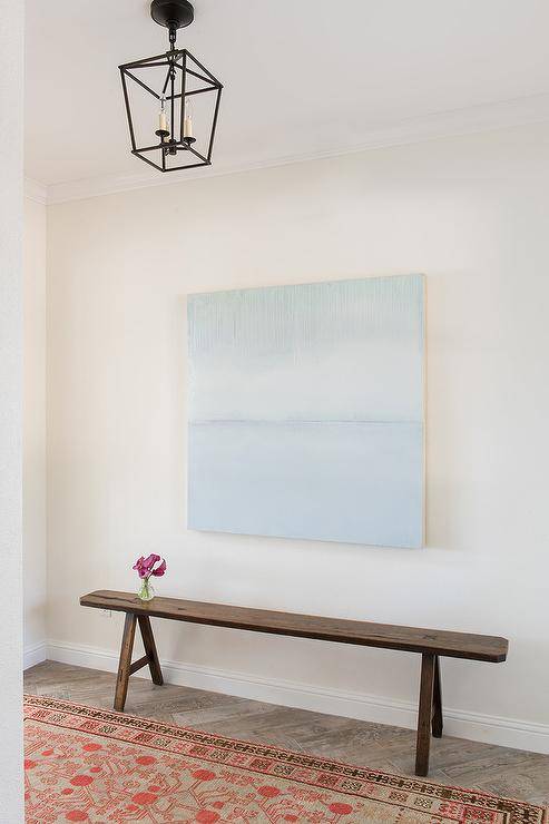 Entryway features blue abstract canvas art over a rustic sawhorse bench and a tan and orange rug, illuminated by a black chandelier.