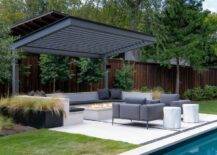 A two-post black pergola is fixed over a built-in u-shaped bench accented with black cushions and pillows and positioned at a concrete fire pit. Gray upholstered accent chairs sit face to face beside an in ground swimming pool.