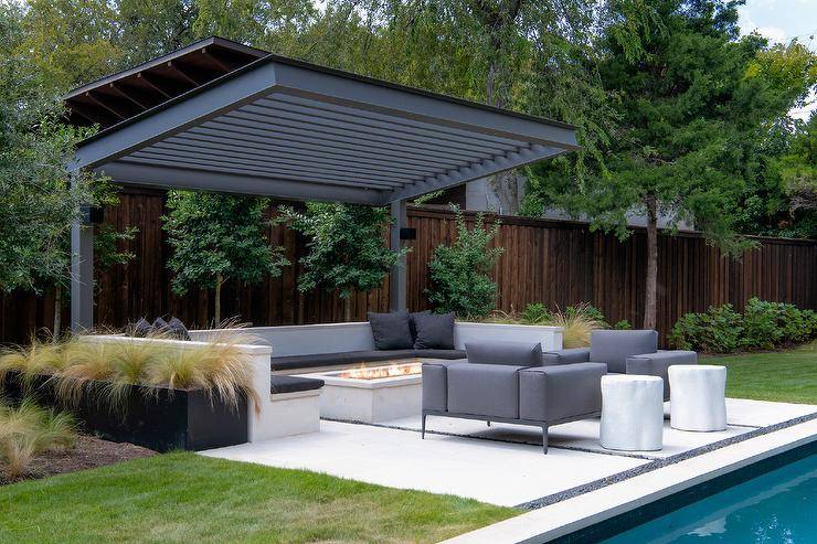 A two-post black pergola is fixed over a built-in u-shaped bench accented with black cushions and pillows and positioned at a concrete fire pit. Gray upholstered accent chairs sit face to face beside an in ground swimming pool.