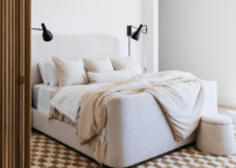 Bedroom features an ivory boucle wingback bed on a brown checkered wool rug, lit by black black sconces.