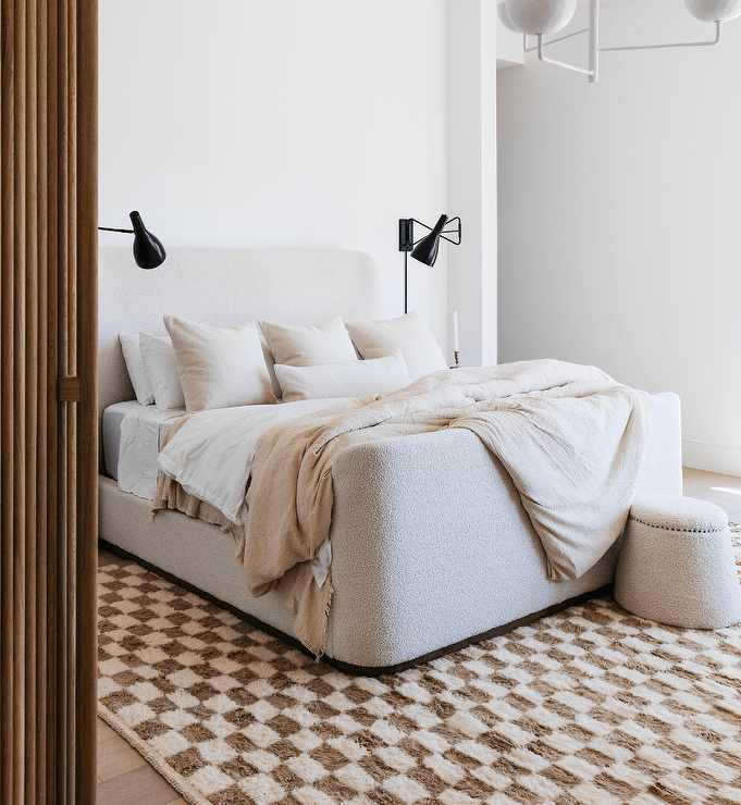 Bedroom features an ivory boucle wingback bed on a brown checkered wool rug, lit by black black sconces.