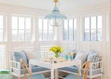 Blue French bistro chairs are placed at a white game table positioned on a painted octagon floor in front of an l-shaped white settee topped with blue cushions and white and blue pillows. The table is illuminated by a blue pagoda lantern and positioned in front of window covered in white cafe shutters.