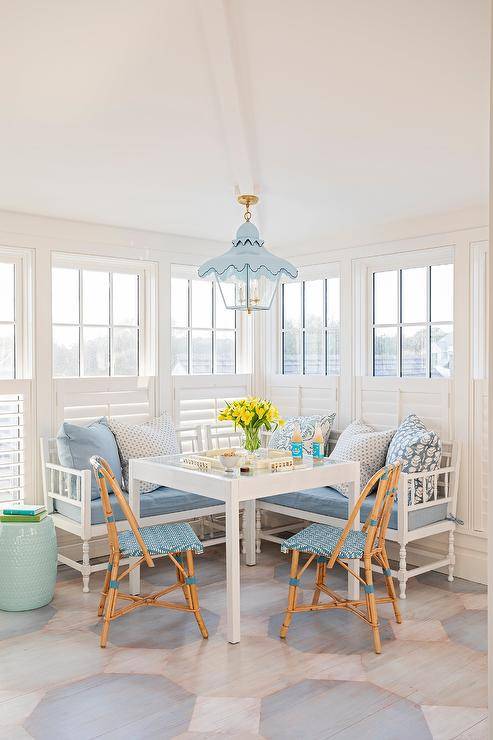 Blue French bistro chairs are placed at a white game table positioned on a painted octagon floor in front of an l-shaped white settee topped with blue cushions and white and blue pillows. The table is illuminated by a blue pagoda lantern and positioned in front of window covered in white cafe shutters.