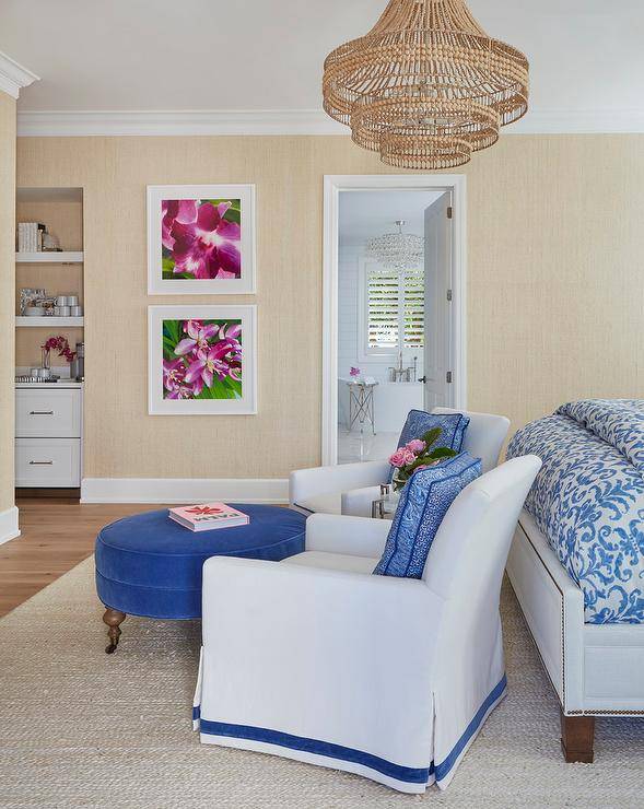 Cottage bedroom design features a sitting area at the foot of the bed with white and blue accent chairs and round blue velvet ottoman illuminated by a rattan cascading chandelier.