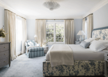 White and blue bedroom boasts a brass floor lamp and a white lattice stool placed beside a blue striped chaise lounge positioned on a blue rug in a corner between windows dressed in ivory curtains. Pale blue walls are complemented with white crown moldings.