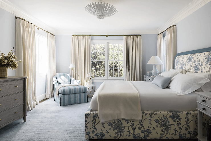 White and blue bedroom boasts a brass floor lamp and a white lattice stool placed beside a blue striped chaise lounge positioned on a blue rug in a corner between windows dressed in ivory curtains. Pale blue walls are complemented with white crown moldings.