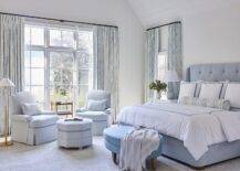 Blue primary bedroom design featrues a blue oval bench at the foot of a tufted bed with blue border shams, a white wooden nightstand lit by a blue lamp and a sitting area with light blue chairs with a light blue octagon ottoman lit by an antique brass floor lamp.