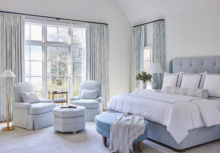 Blue primary bedroom design featrues a blue oval bench at the foot of a tufted bed with blue border shams, a white wooden nightstand lit by a blue lamp and a sitting area with light blue chairs with a light blue octagon ottoman lit by an antique brass floor lamp.