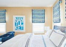 Tan wallpaper covers the walls of a white, blue, and tan bedroom featuring windows covered in tan and blue geometric roman shades flanking white and blue typography art.