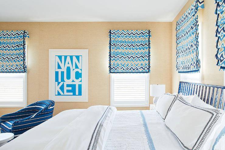 Tan wallpaper covers the walls of a white, blue, and tan bedroom featuring windows covered in tan and blue geometric roman shades flanking white and blue typography art.