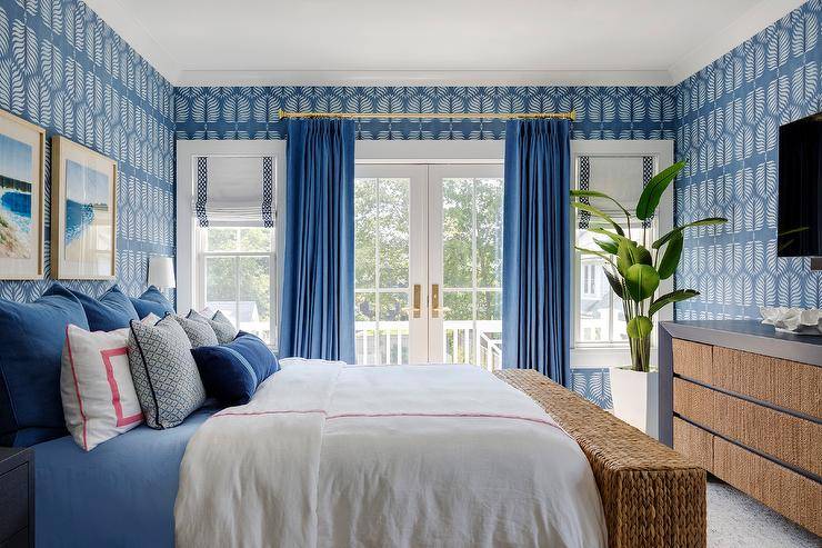 Restful blue bedroom features a bed dressed in red and blue bedding complemented with layered red and blue pillows. A seagrass bench sits at the foot of the bed and in front of a blue wooden dresser positioned under a TV hung from a wall covered in blue botanical wallpaper. Art hangs over the bed, while windows covered in white and blue roman shades flank French doors accented with blue curtains.