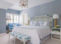 Positioned against a wall clad in blue grasscloth wallpaper, a headboard in Clarence House Tibet fabric supports a bed dressed in white and blue bedding topped blue monogrammed shams. A white bamboo bench sits at the foot of the bed, while the bed is flanked by white nightstands lit by gold lamps.