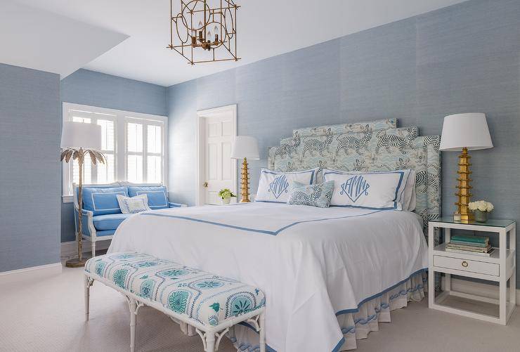 Positioned against a wall clad in blue grasscloth wallpaper, a headboard in Clarence House Tibet fabric supports a bed dressed in white and blue bedding topped blue monogrammed shams. A white bamboo bench sits at the foot of the bed, while the bed is flanked by white nightstands lit by gold lamps.