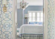 Blue bedroom walls are lit by a silver leaf floor lamp and a brass lantern hung in front of a bed covered in white and blue bedding with a white and blue bedskirt.