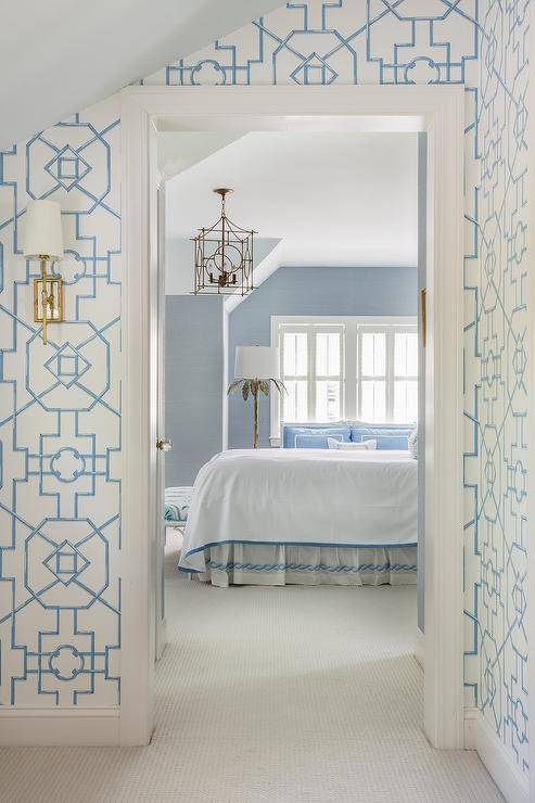 Blue bedroom walls are lit by a silver leaf floor lamp and a brass lantern hung in front of a bed covered in white and blue bedding with a white and blue bedskirt.