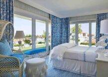 White and blue bedroom features a white and blue bamboo bench in front large glass paneled doors with blue curtains, a bed with blue and white bedding, a cream bedside table lit by a white and blue floral lamp and a large rope and rattan accent chair with blue cushions and a white tassel stool atop a blue rug.