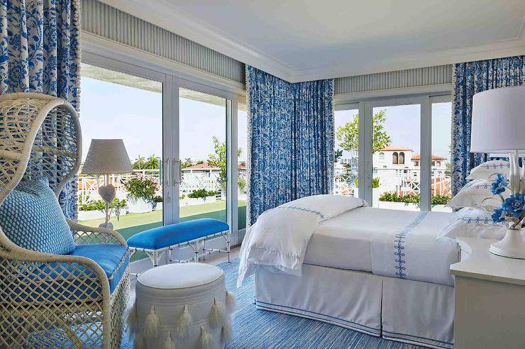 White and blue bedroom features a white and blue bamboo bench in front large glass paneled doors with blue curtains, a bed with blue and white bedding, a cream bedside table lit by a white and blue floral lamp and a large rope and rattan accent chair with blue cushions and a white tassel stool atop a blue rug.