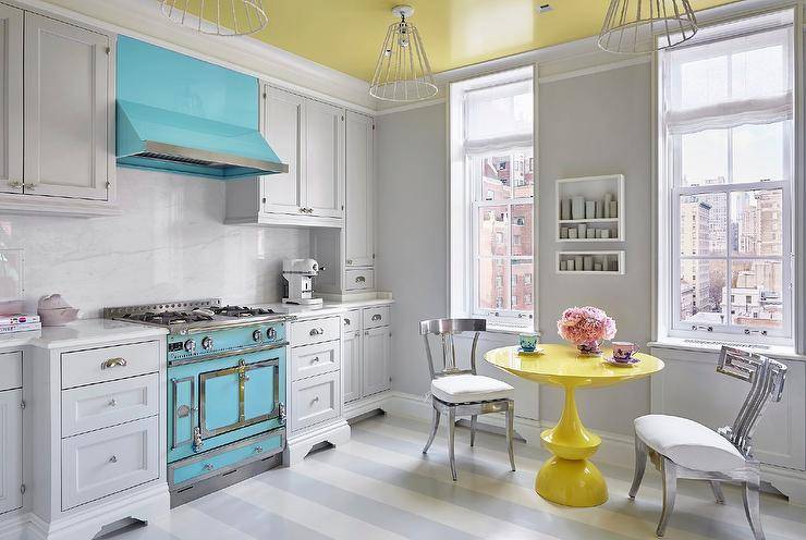 A colorful yellow, blue, and gray kitchen showcases a bright yellow lacquer dining table flanked by silver klismos chair placed on a white and gray stripe painted floor. Above the table, white shelves are mounted beneath a yellow painted ceiling to a gray wall and stacked between windows. A turquoise blue stove sits between light gray shaker drawers accented with polished nickel hardware and a white quartz countertop fitted against a marble slab backsplash. While above the stove, a turquoise blue range hood is located between light gray shaker upper cabinets.