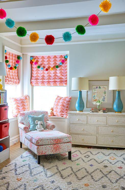 Fun and colorful girls bedroom boasting lots of colors and bold patterns. This bright room is designed with an ivory bone inlay dresser displaying two tall turquoise blue lamps with wall art and styled decor in between. A white and pink chaise lounge is furnished in the corner of the bedroom under two windows fitted with pink and orange geometric roman shades. Details that show off tons of personality and mix the colors and patterns are pom pom garland and colorful shag area rug.