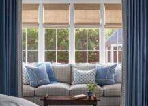 Bamboo roman shades cover a row of windows framed by a blue wall and positioned above a gray grid couch complemented with blue pillows. The sofa is paired with an antique French coffee table, while the sitting area is located behind blue curtains.