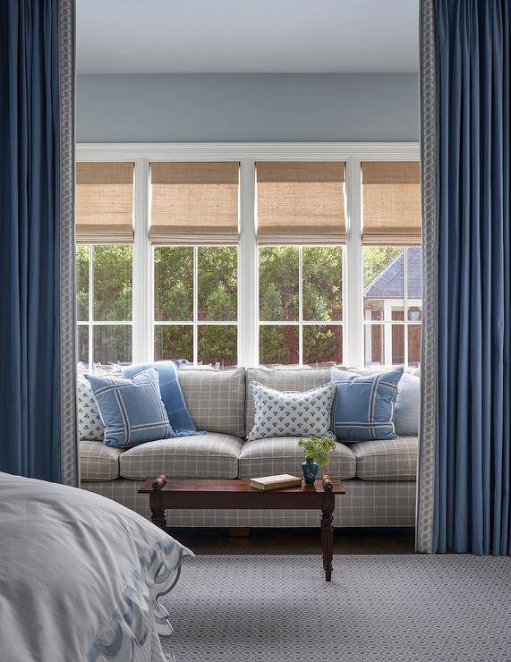 Bamboo roman shades cover a row of windows framed by a blue wall and positioned above a gray grid couch complemented with blue pillows. The sofa is paired with an antique French coffee table, while the sitting area is located behind blue curtains.